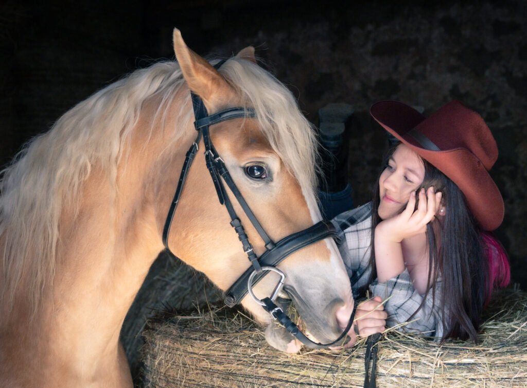 Haflinger daneben junges Mädchen auf einem Strohballen liegend mit rotem Cowboyhut